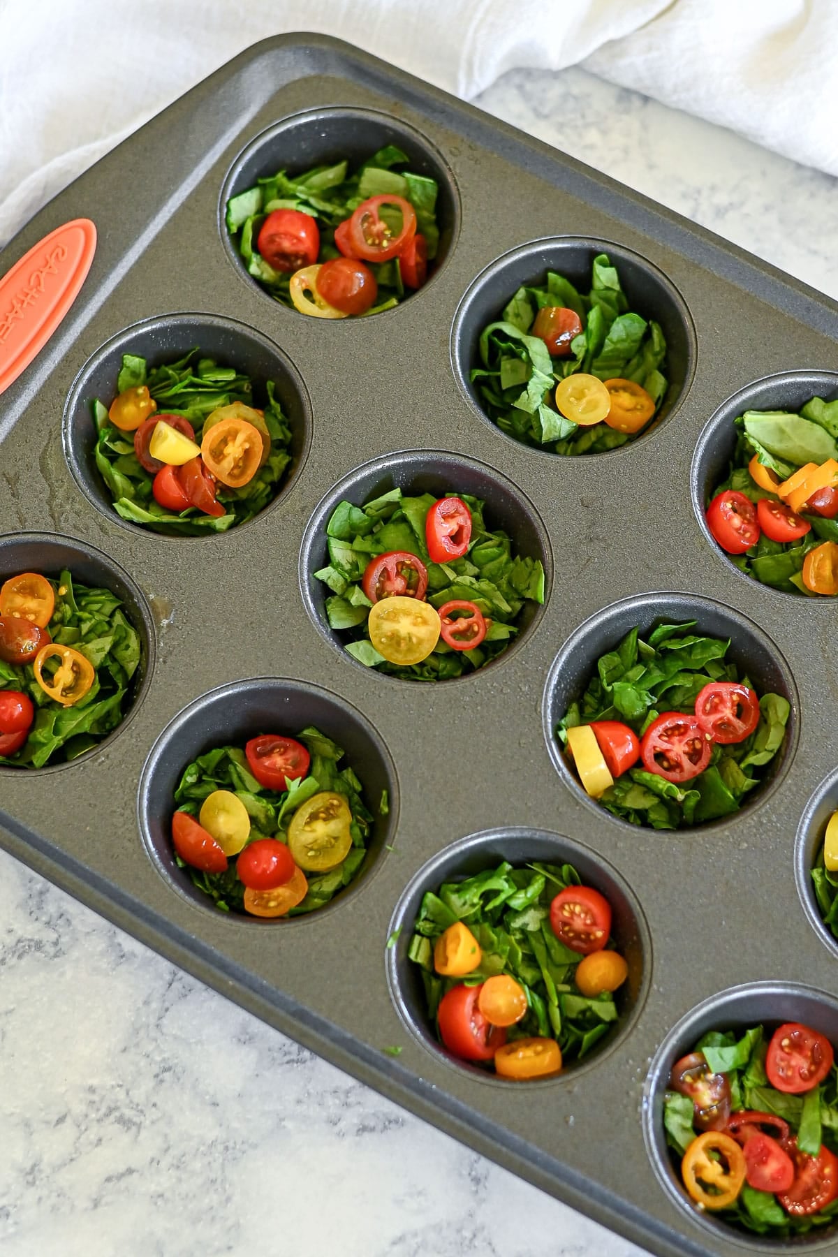 A muffin tin filled with chopped spinach and halved cherry tomatoes on a marble countertop, perfect for prepping Spinach Egg Cups.