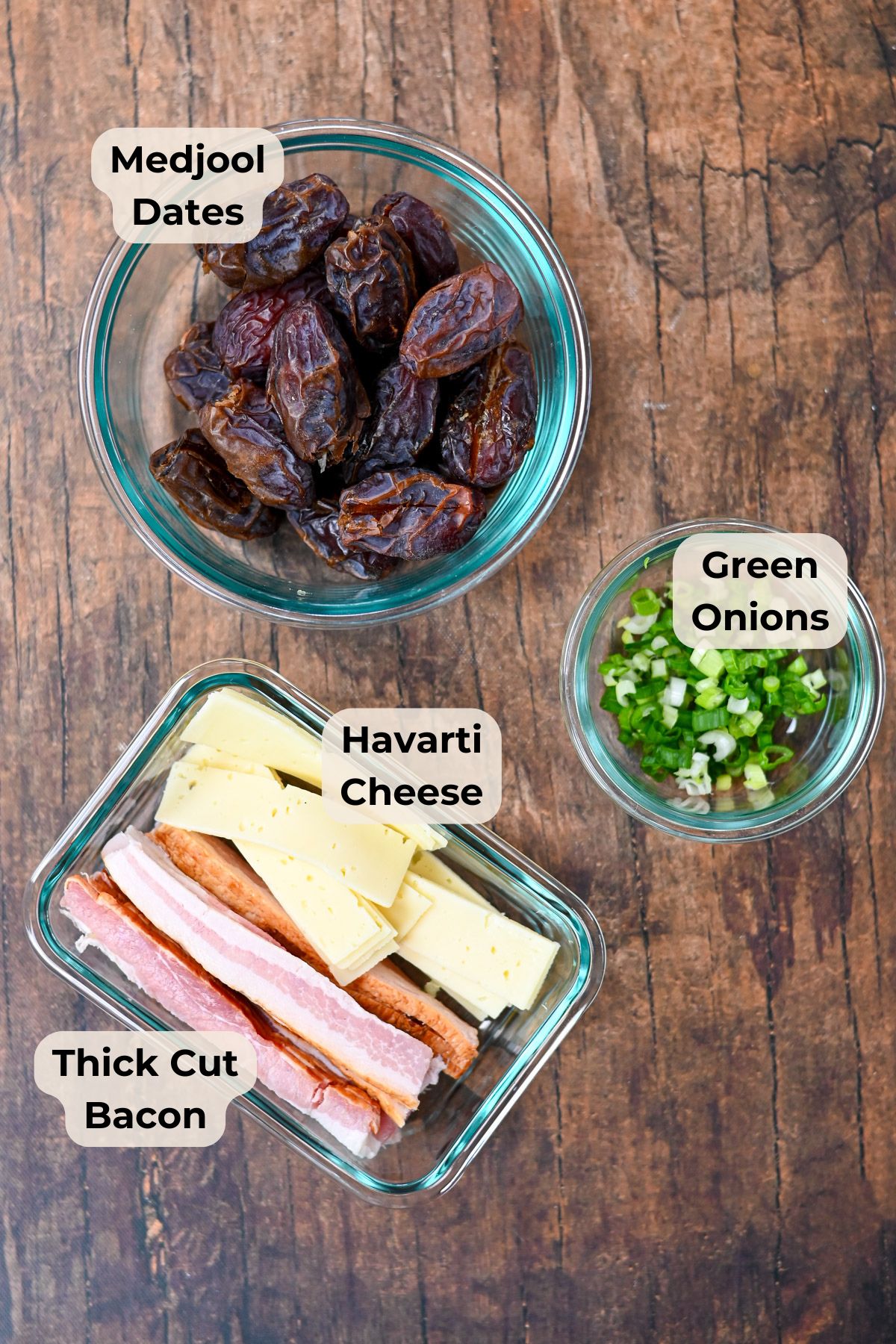 Three bowls with labeled ingredients-Medjool dates, green onions, Havarti cheese, and thick cut bacon-sit on a wooden table, ready to make a delicious appetizer: Cheese Stuffed Dates Wrapped in Bacon.