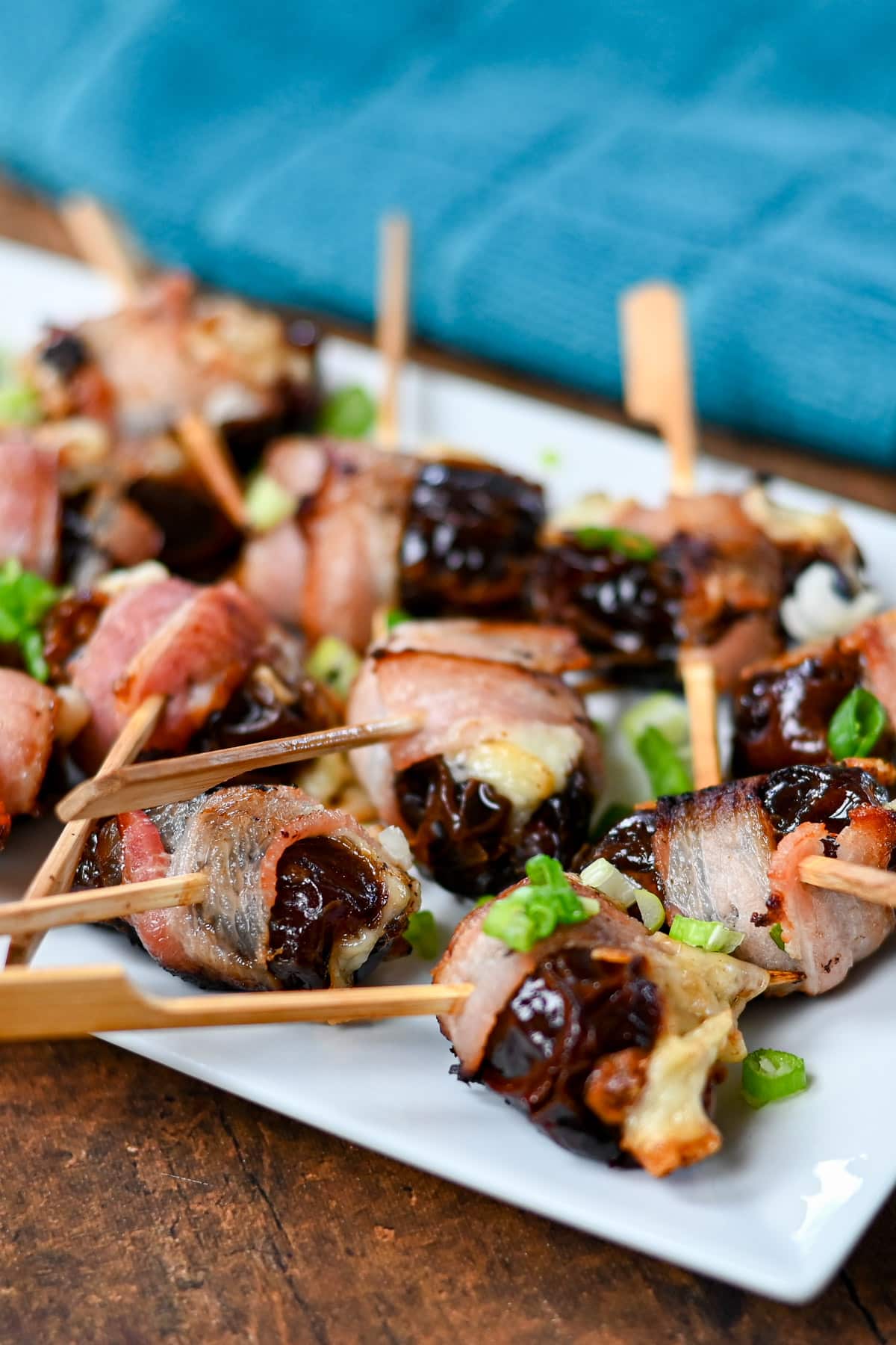 Cheese Stuffed Dates Wrapped in Bacon are skewered and garnished with chopped green onions on a white plate.