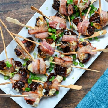Cheese Stuffed Dates Wrapped in Bacon are skewered and garnished with chopped green onions, making a savory appetizer beautifully presented on a white platter.