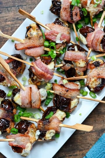 Cheese Stuffed Dates Wrapped in Bacon are skewered and garnished with chopped green onions, making a savory appetizer beautifully presented on a white platter.