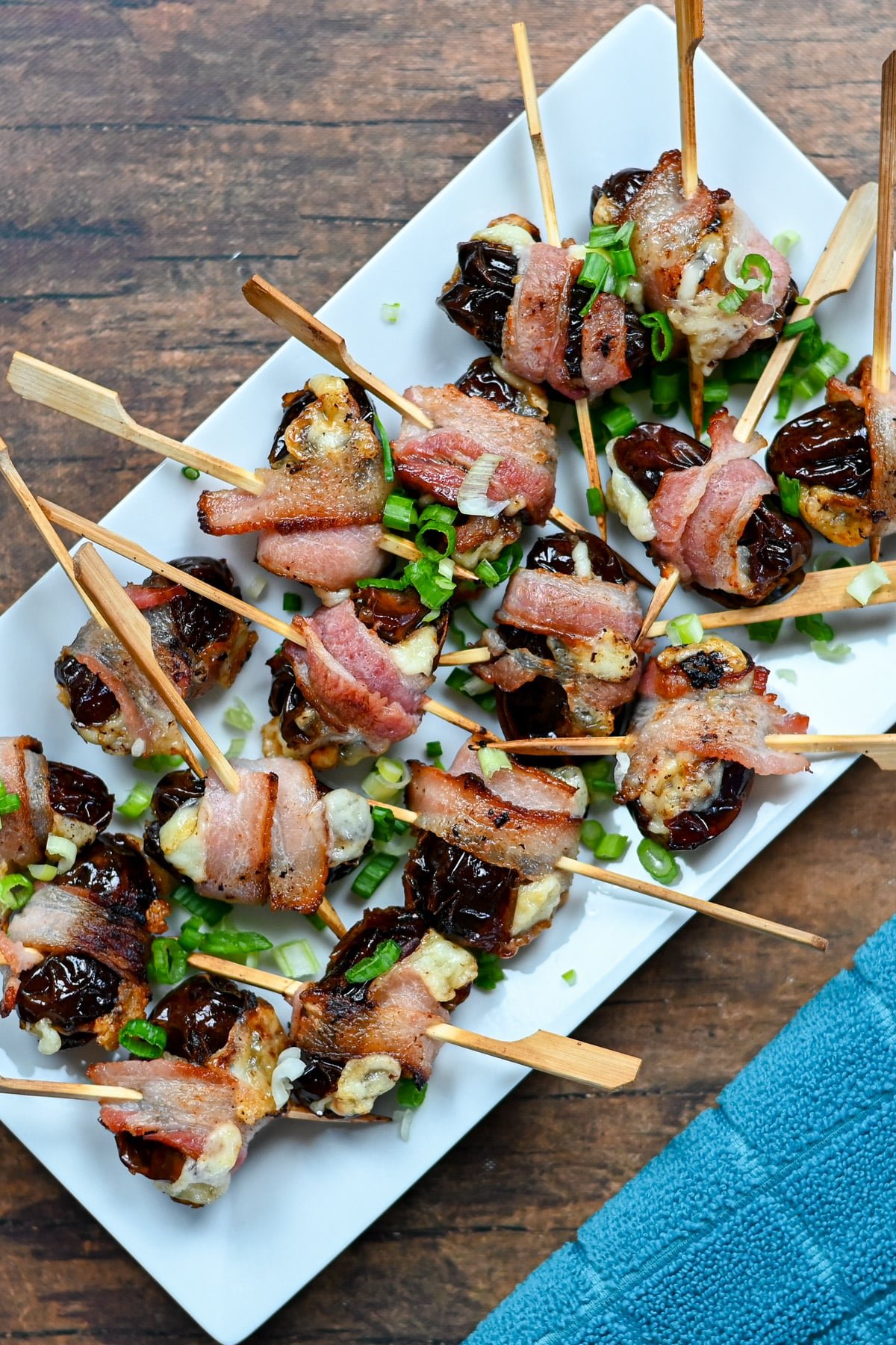 Cheese Stuffed Dates Wrapped in Bacon on skewers, served as an irresistible appetizer on a white plate and garnished with chopped green onions.