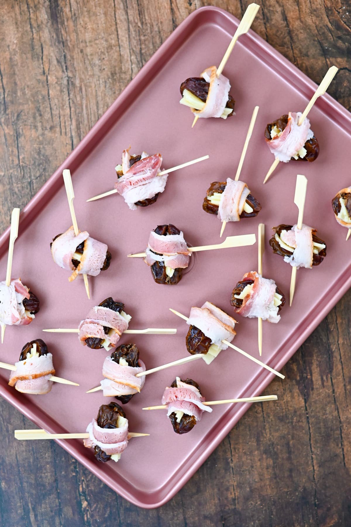 Cheese Stuffed Dates Wrapped in Bacon and nuts, served on skewers atop a pink baking tray, make the perfect appetizer for any occasion.