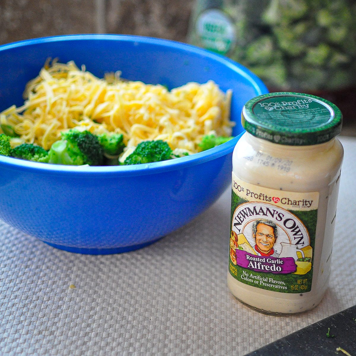 a jar of Newman's Own alfredo sauce next to a blue bowl of chicken, broccoli, and shredded cheese. 