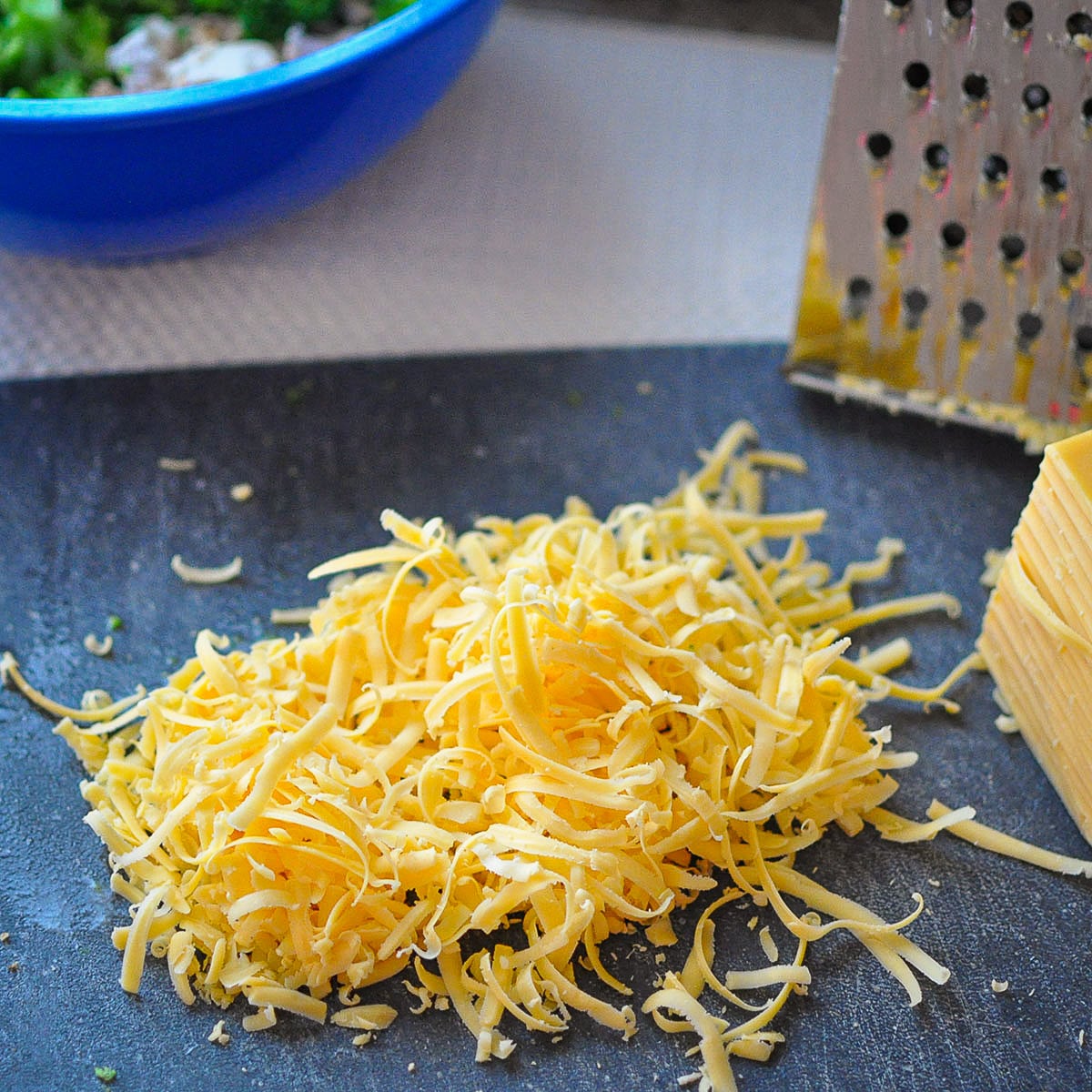 a pile of shredded cheddar cheese on a black cutting board next to a box grater. 