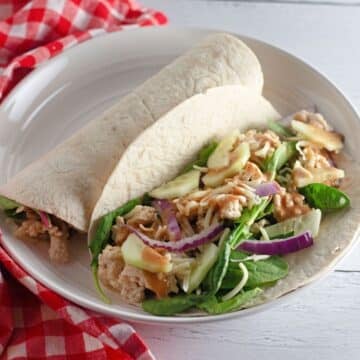 A tortilla wrap filled with spinach, chicken, red onion, cheese, and sauce on a white plate-try our Chicken Sesame Ginger Wraps for a tasty twist.