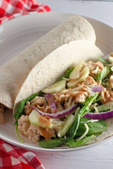 A tortilla wrap filled with spinach, chicken, red onion, cheese, and sauce on a white plate-try our Chicken Sesame Ginger Wraps for a tasty twist.