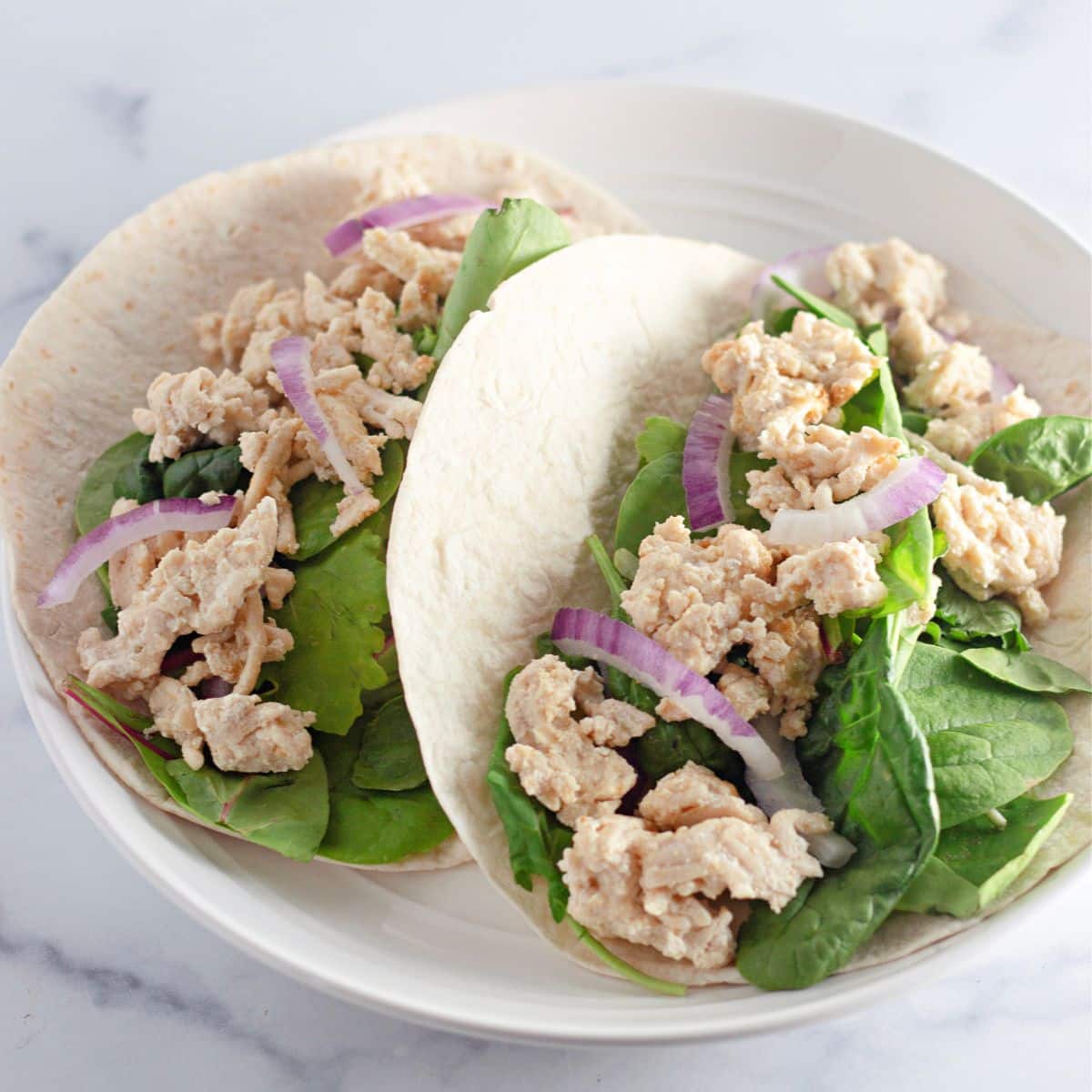 Chicken Sesame Ginger Wraps: Two soft tortillas filled with leafy greens, shredded chicken, and sliced red onions on a white plate.