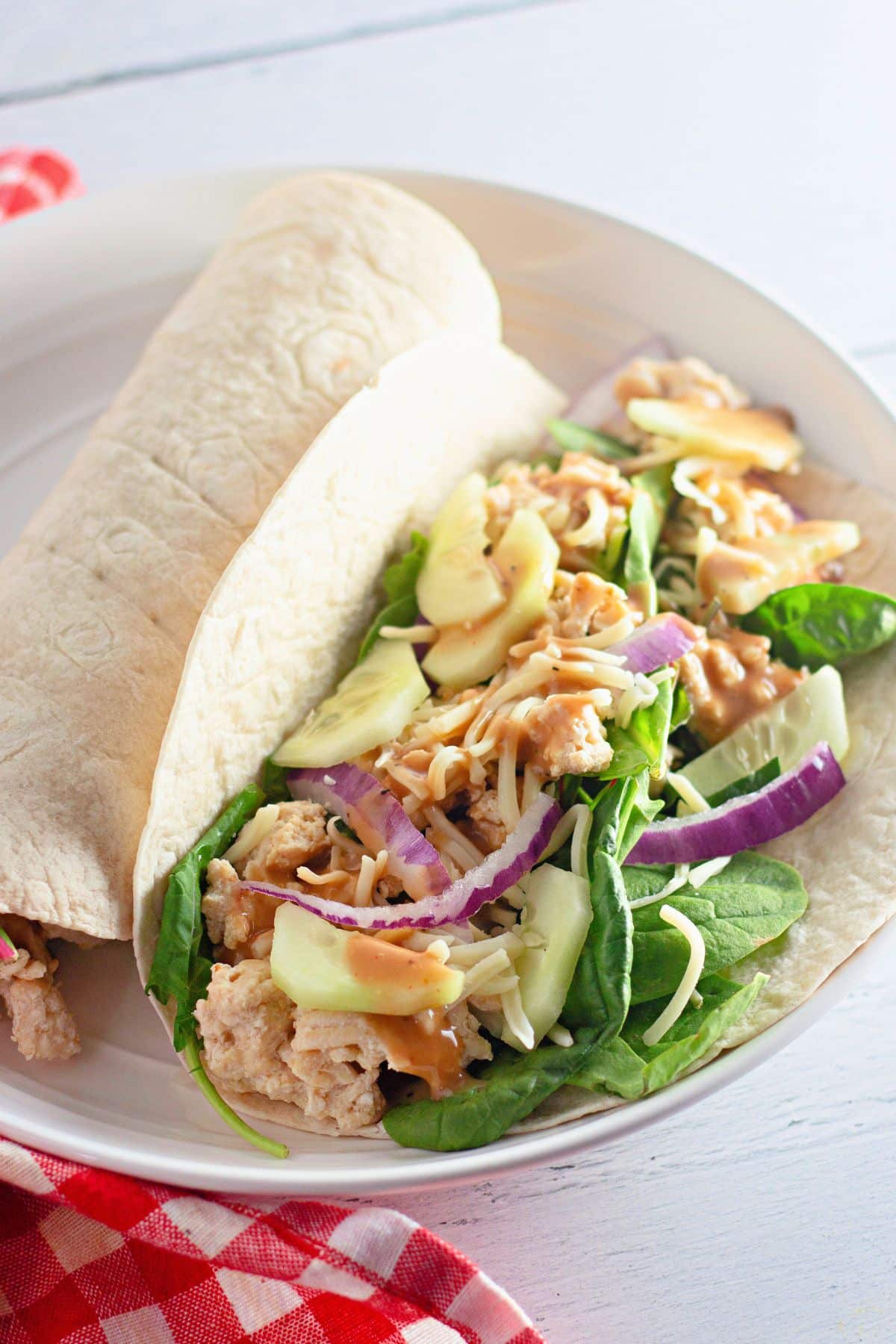 Two Chicken Sesame Ginger Wraps filled with spinach, red onion, cheese, apple slices, and a drizzle of sauce on a white plate.