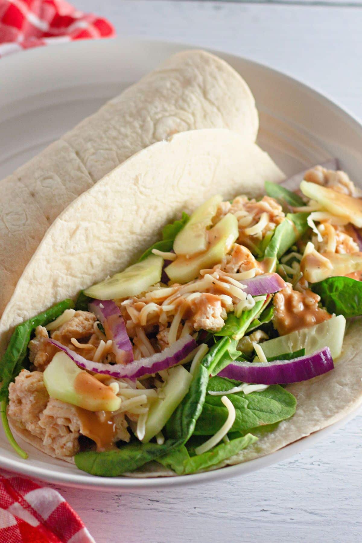 Sesame Ginger Wraps-a tortilla filled with tender chicken, spinach, sliced apples, red onions, cheese, and a flavorful dressing, all served on a white plate.