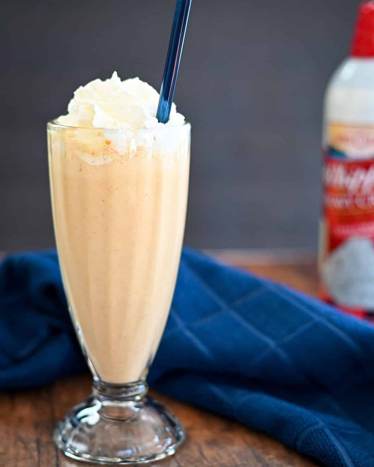 Milkshake in a glass with a blue straw next to a blue towel and a can of whipped topping.
