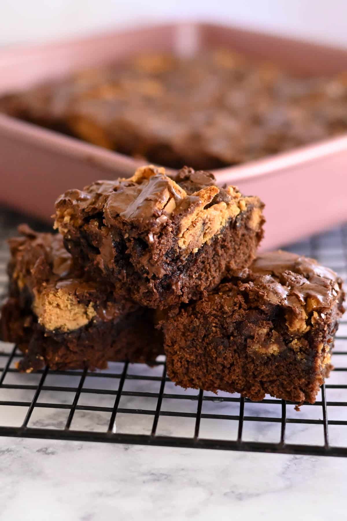 A stack of Nutter Butter Chocolate Chip Brownies with a creamy peanut butter filling rests on a cooling rack, tempting with their rich aroma, while another pan of these mouthwatering treats sits invitingly in the background.