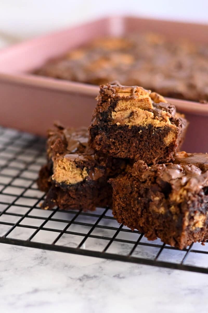 Nutter Butter Chocolate Chip Brownies stacked on a cooling rack, showcasing tantalizing brownie squares topped with cookie pieces. A pan lingers in the background, hinting at more treats to come.