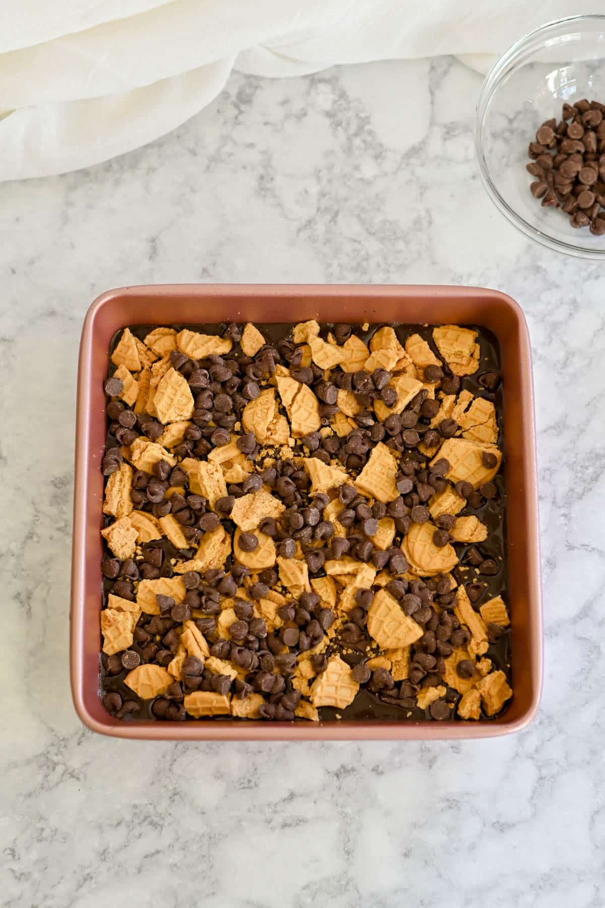 A baking pan brimming with Nutter Butter Chocolate Chip Brownies rests on the cool marble countertop, surrounded by a bowl of chocolate chips. 
