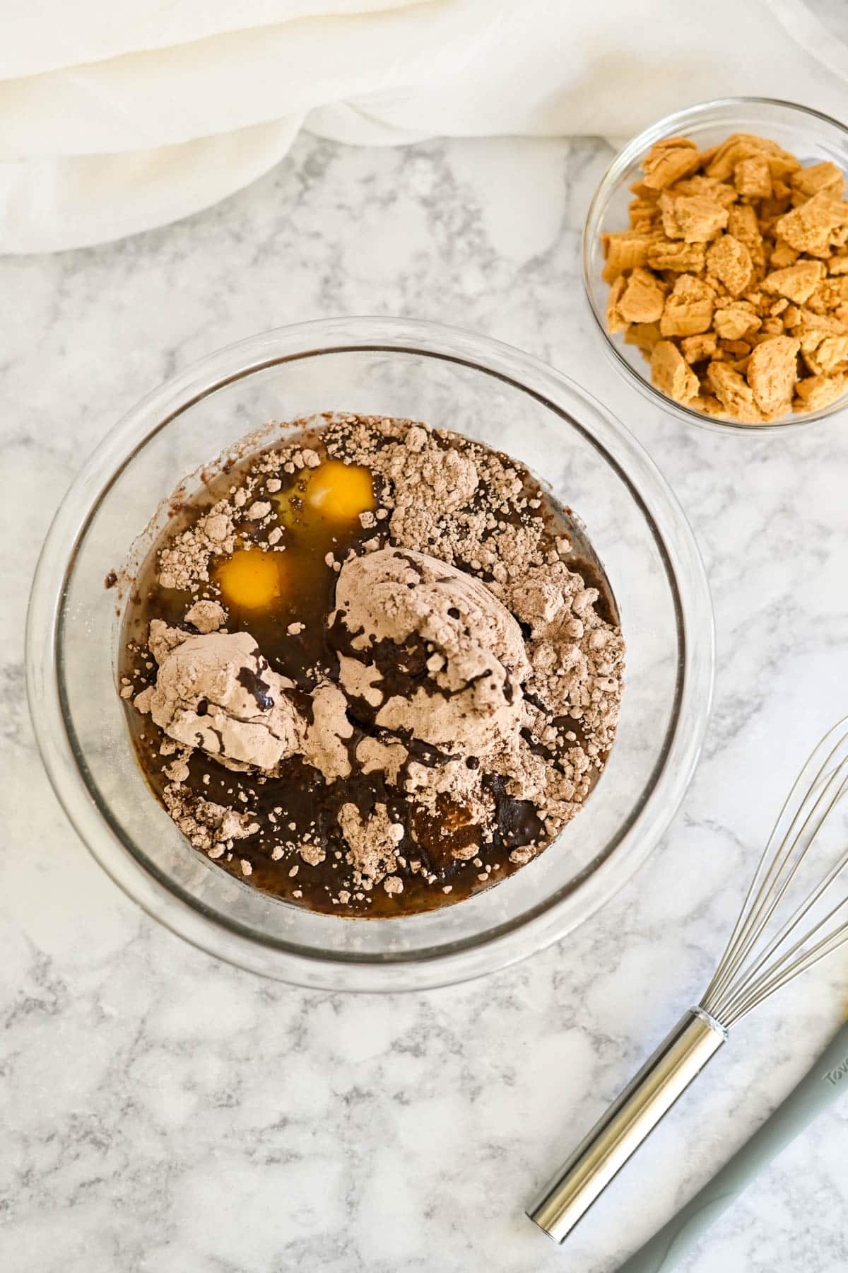 A mixing bowl with eggs, chocolate powder, liquid, and a whisk sits beside a cereal-filled bowl on the marble surface.