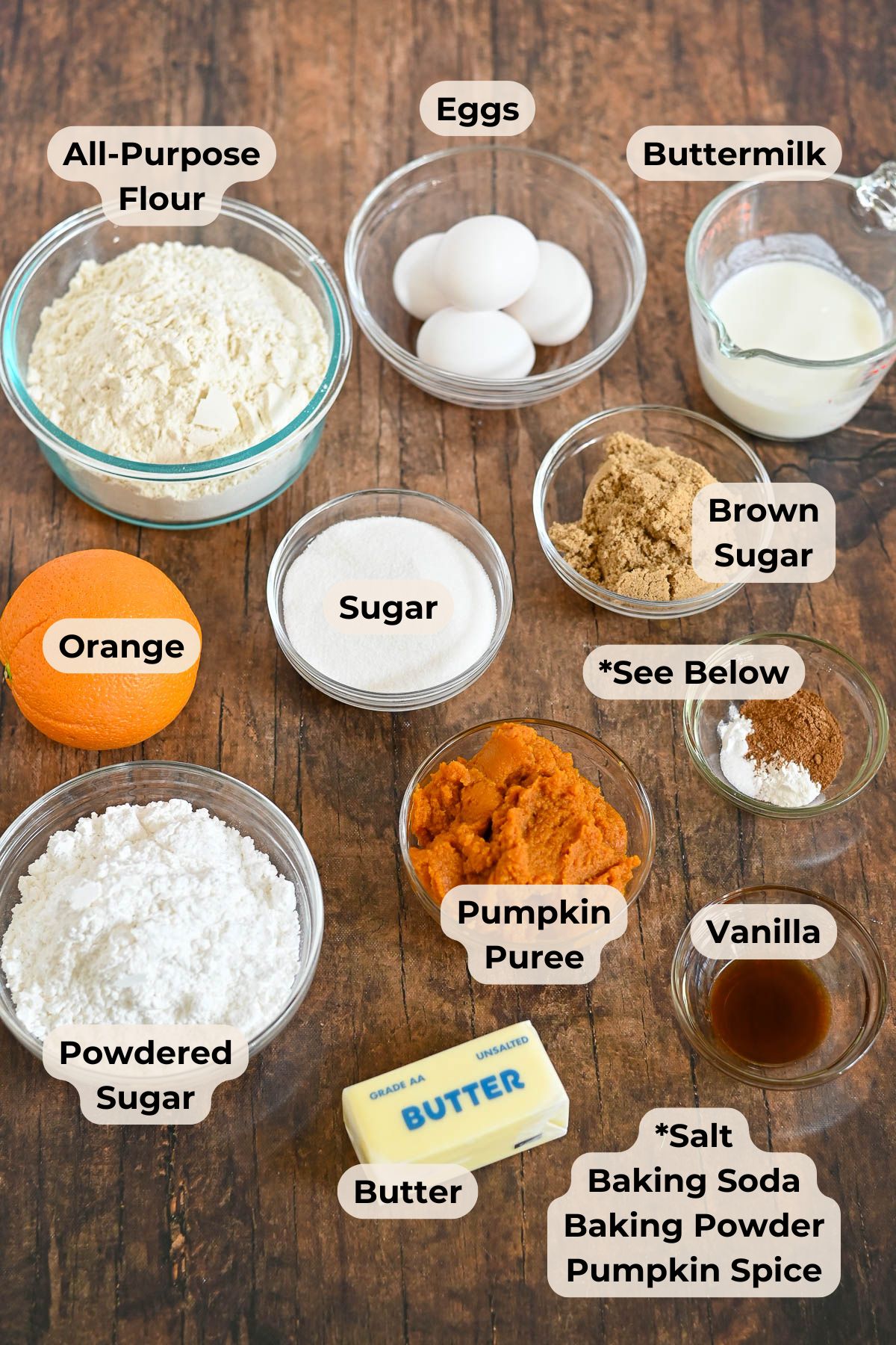 Various labeled baking ingredients in bowls on a wooden table, including eggs, flour, sugars, pumpkin puree, butter, and spices-everything you need to create a delicious Pumpkin Bundt Cake.