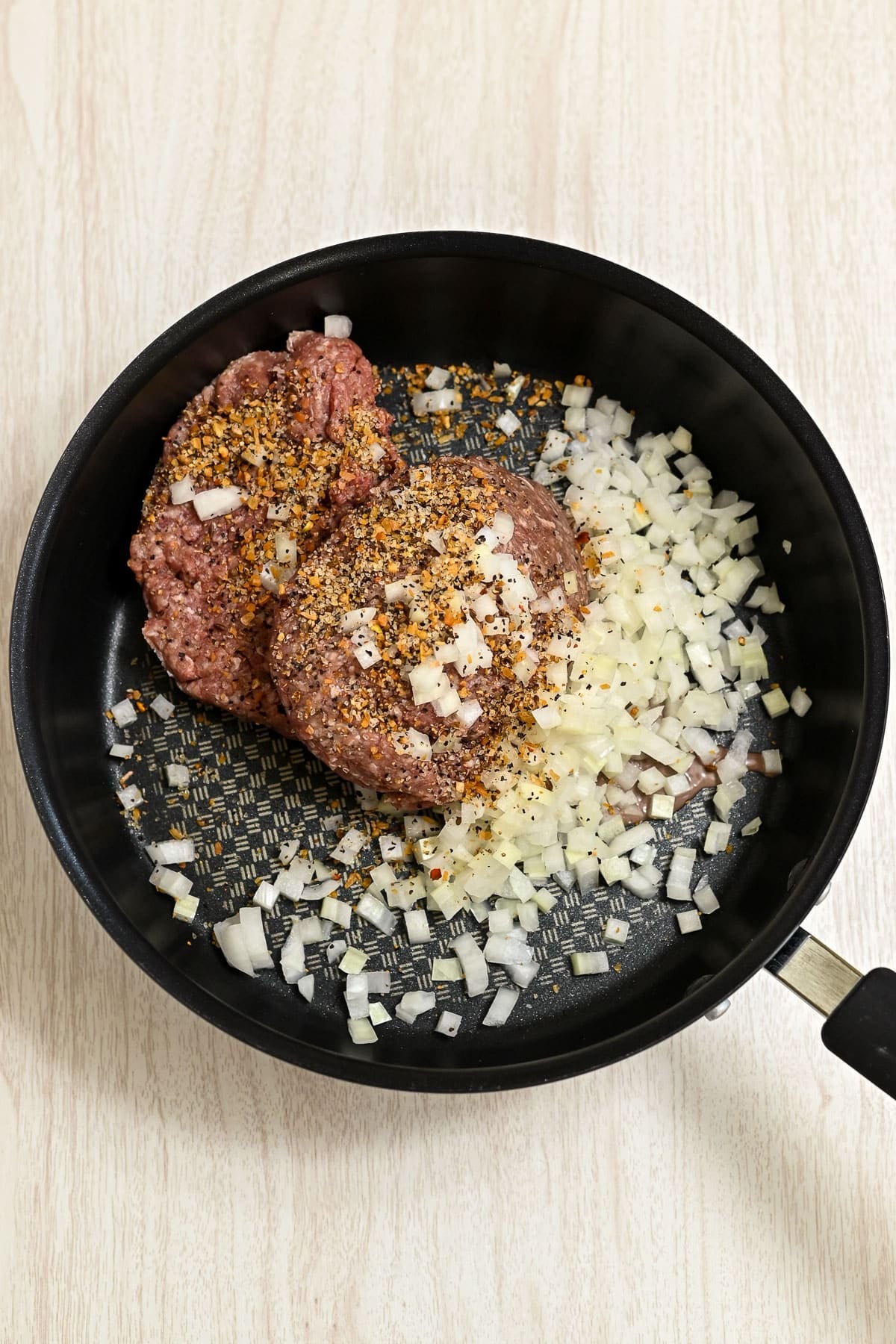 Ground beef with chopped onions and spices in a black frying pan on a light wooden surface.