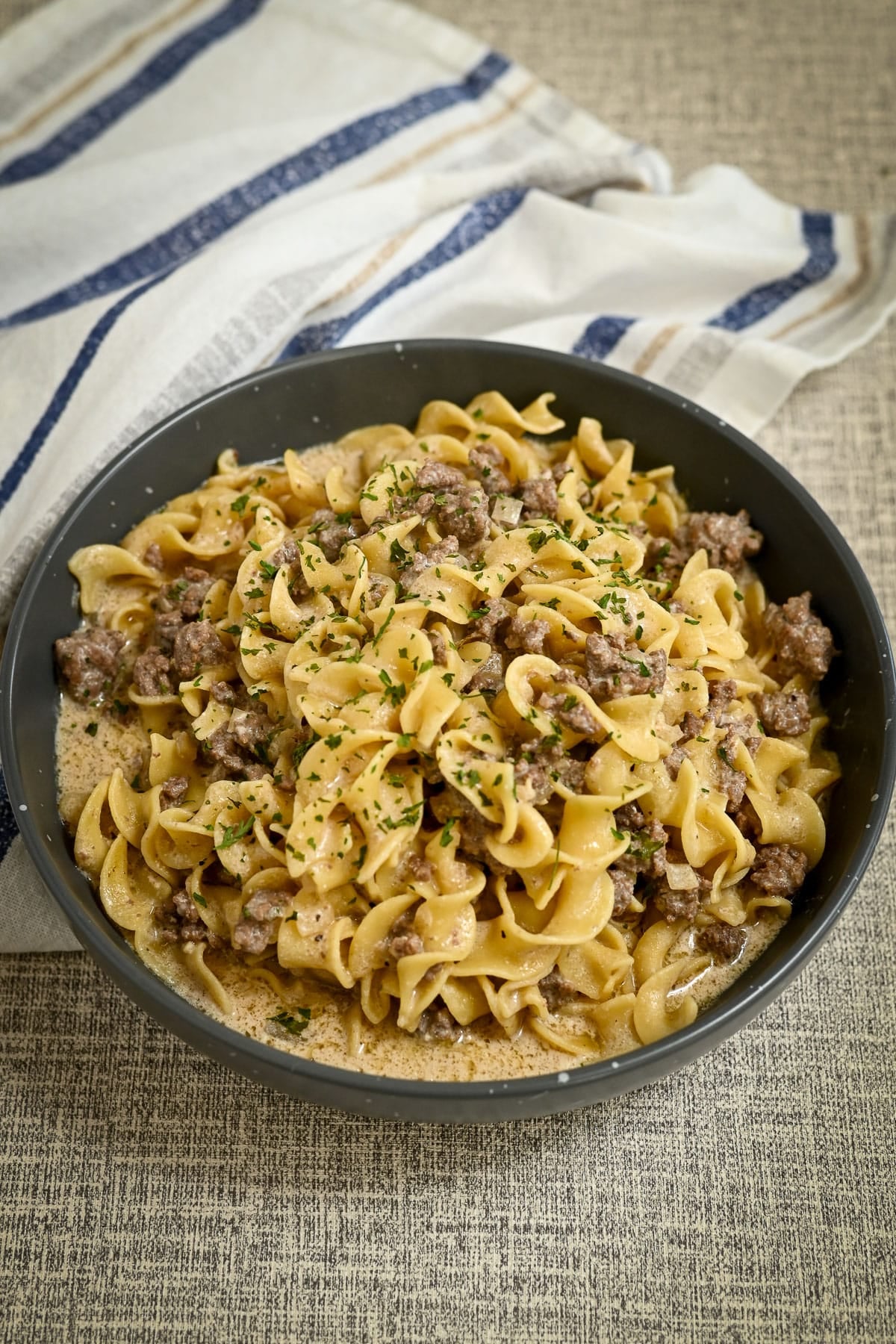 A bowl of creamy ground beef and noodle pasta, reminiscent of a Simple Hamburger Stroganoff, garnished with chopped herbs and served on a table with a striped towel.