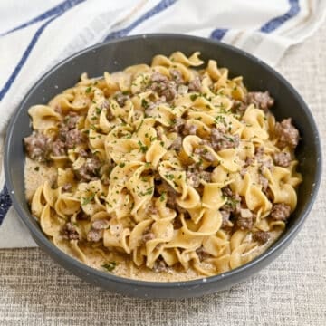 A bowl of creamy ground beef stroganoff with egg noodles, topped with chopped parsley, on a textured surface. This simple hamburger stroganoff makes for a comforting and delicious meal.