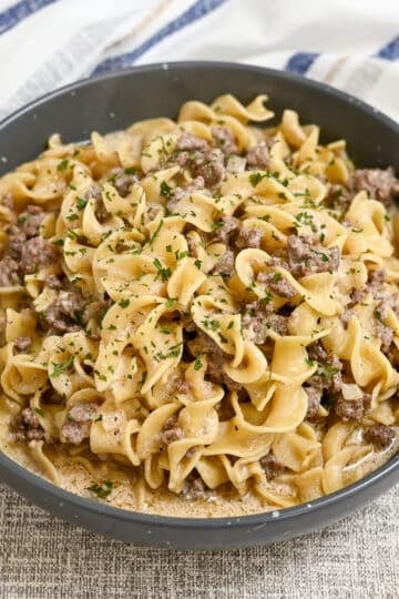 A bowl of creamy ground beef stroganoff with egg noodles, topped with chopped parsley, on a textured surface. This simple hamburger stroganoff makes for a comforting and delicious meal.