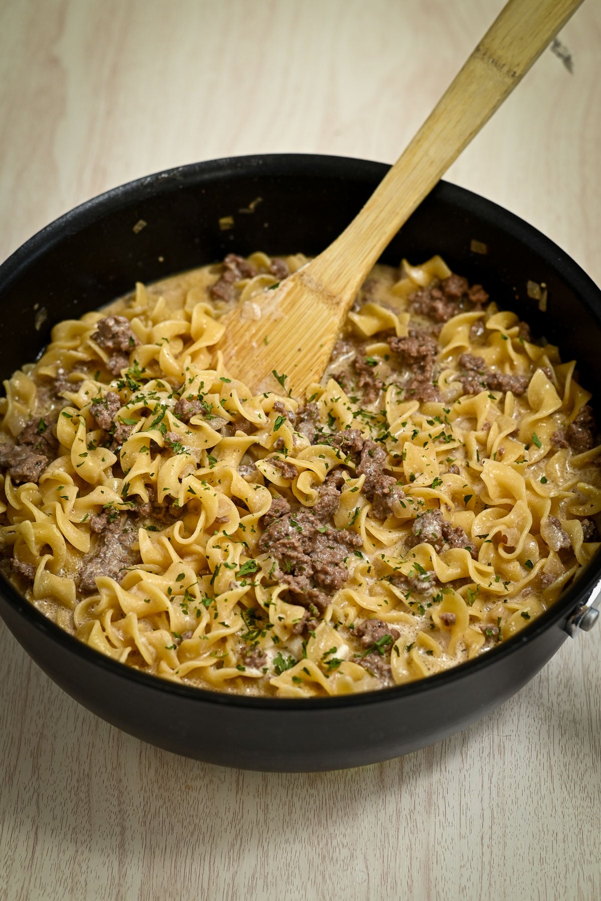 Wide skillet of Simple Hamburger Stroganoff made with ground beef and egg noodles, garnished with herbs, and a wooden spoon for serving.