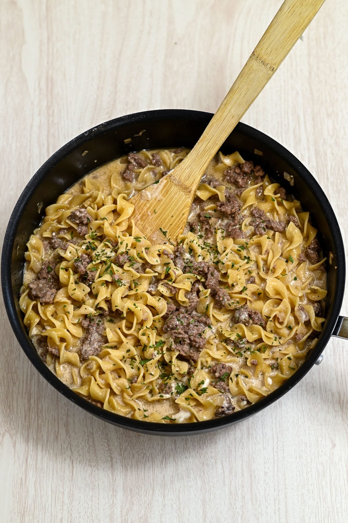 A skillet of creamy Simple Hamburger Stroganoff, made with ground beef and noodles, sits with a wooden spoon on a light wooden surface.