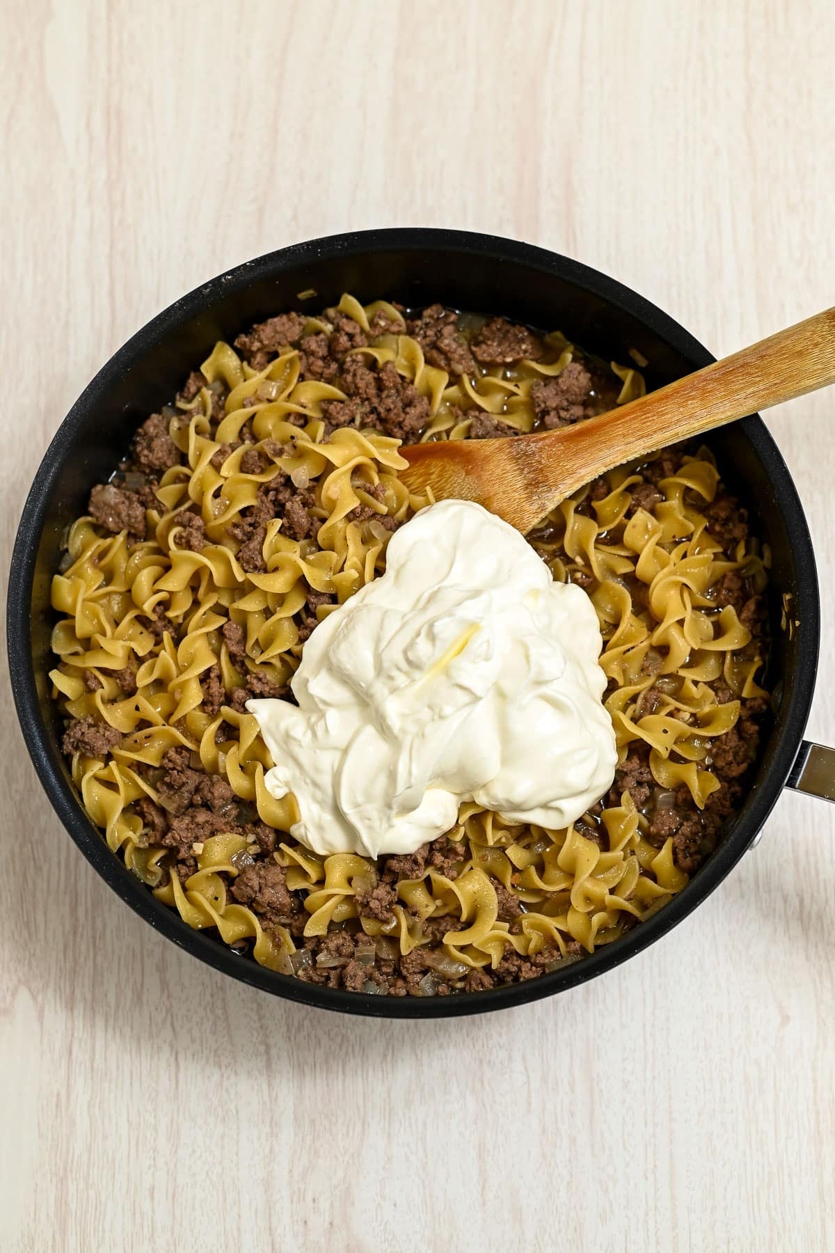 A skillet with cooked noodles, ground beef, and a large dollop of sour cream being stirred with a wooden spoon.