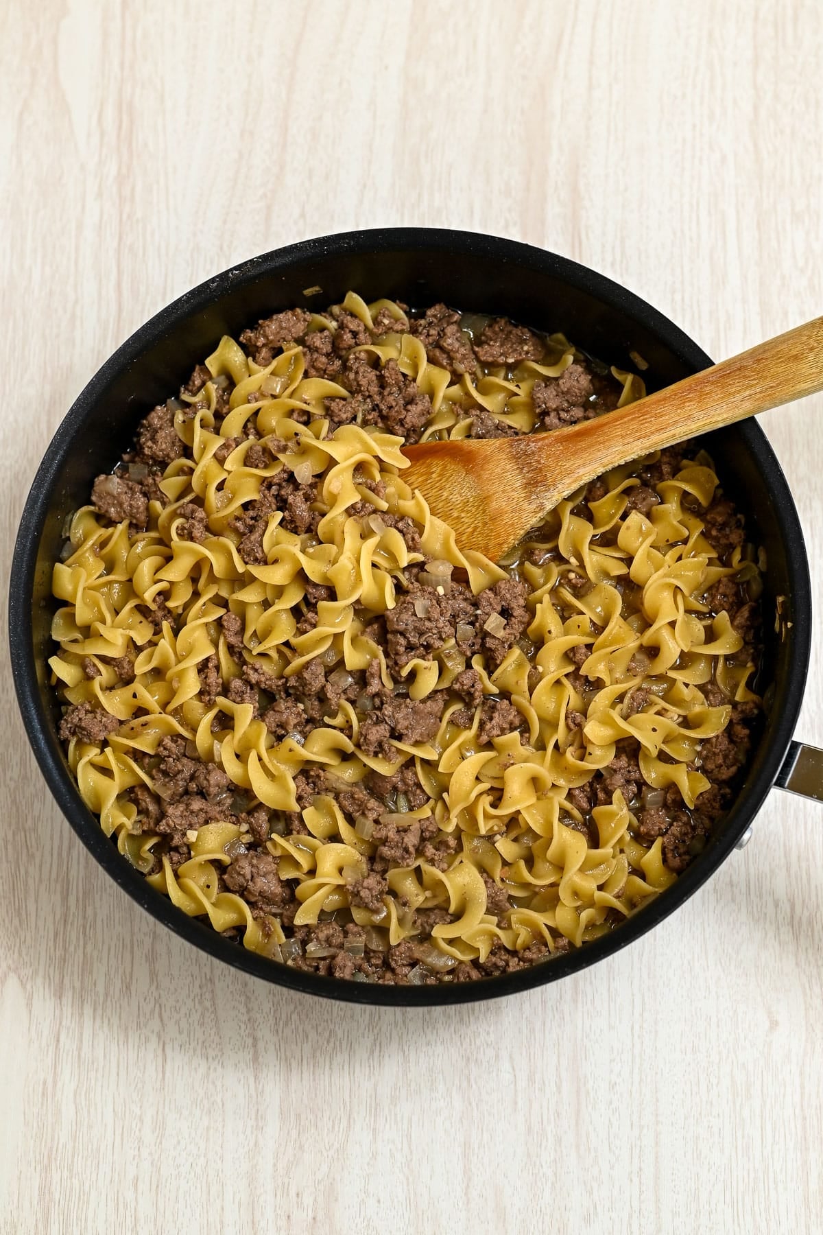 A skillet of cooked egg noodles and ground beef, perfect for a Simple Hamburger Stroganoff, with a wooden spoon on a light wood surface.