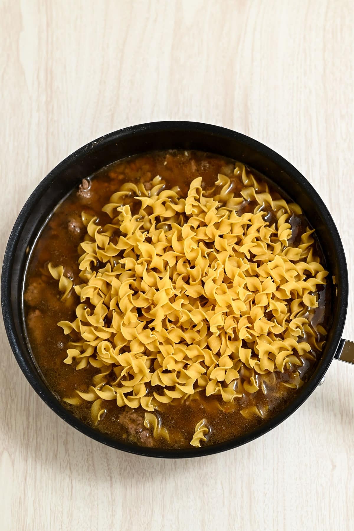 Uncooked egg noodles in a skillet with ground beef and broth on a light wood surface.