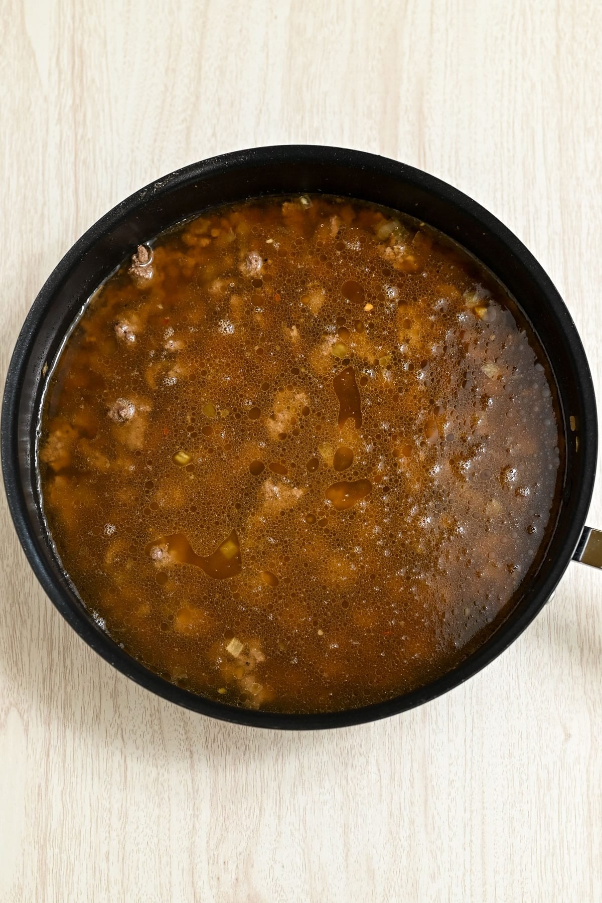 A pot filled with beef broth and ground beef, simmering on a light wooden surface.