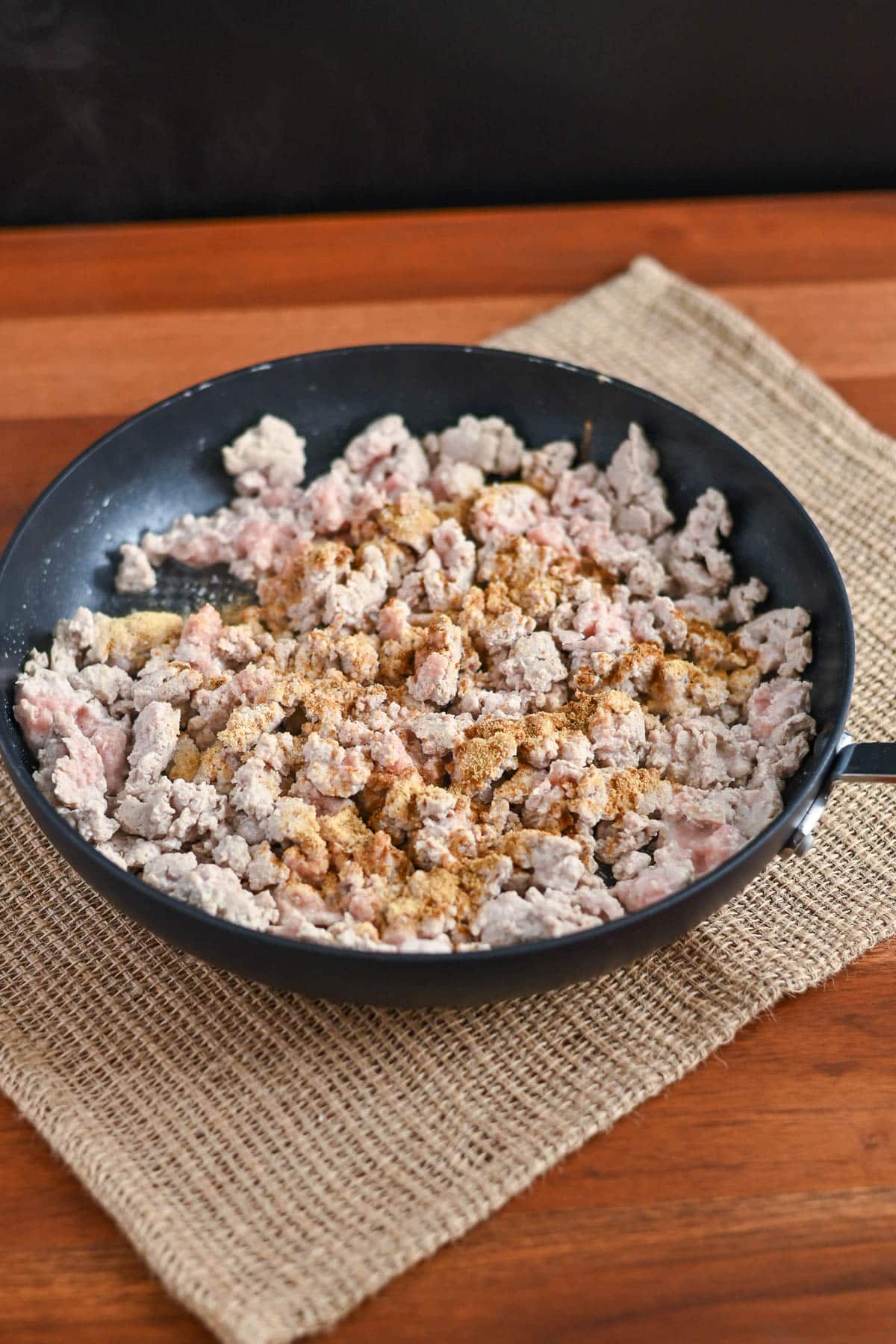 Ground turkey cooking in a black skillet, sprinkled with seasoning, on a burlap mat and wooden surface-perfect for Taco Sloppy Joes recipes.