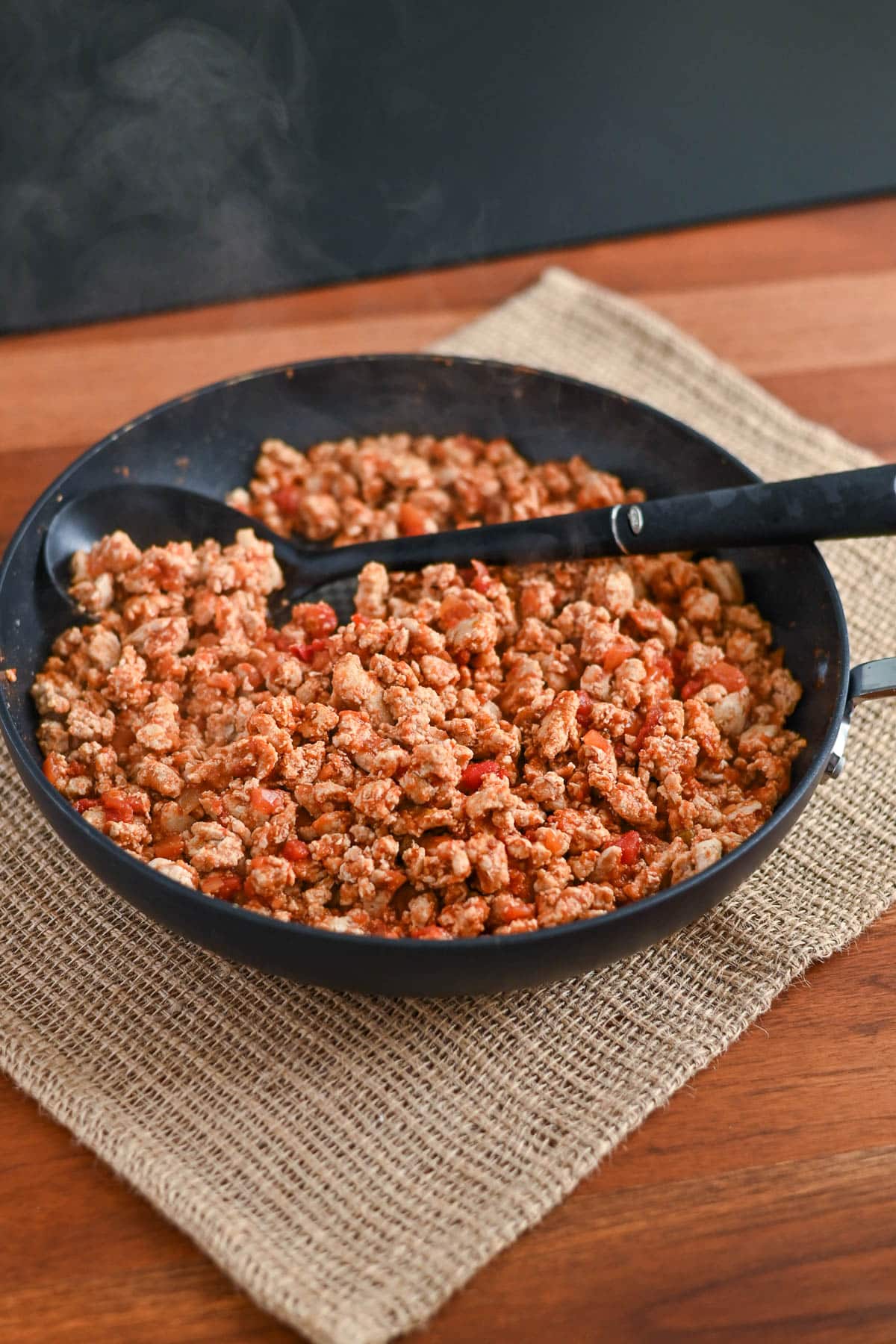 A skillet filled with cooked ground turkey and diced tomatoes, perfect for Taco Sloppy Joes recipes, sits on a woven mat with two black utensils.