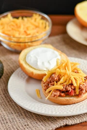 Open sandwich bun with ground meat, sour cream, and shredded cheese on a plate; Taco Sloppy Joes recipes ingredients in background.