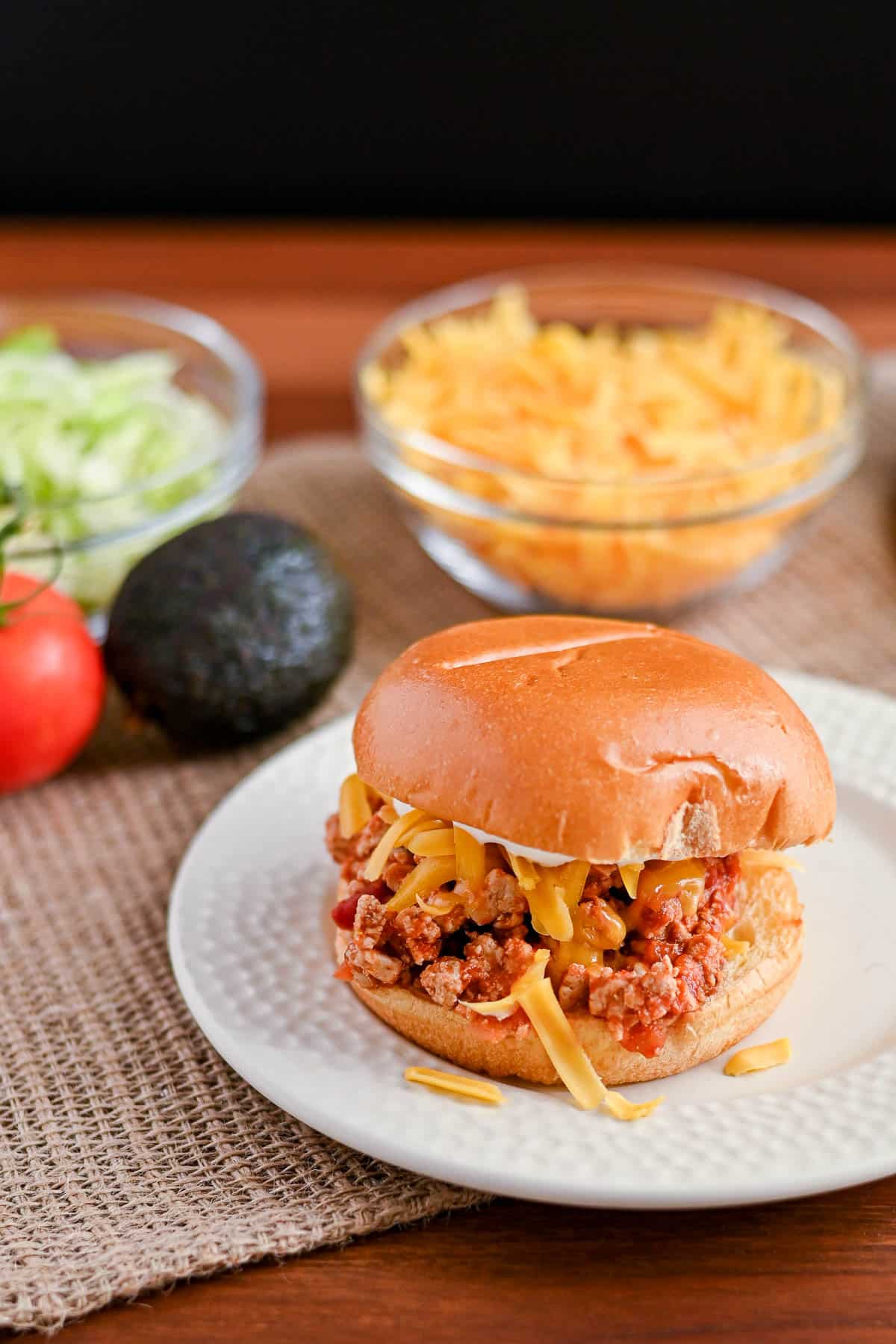 A Taco Sloppy Joes sandwich with shredded cheese on a plate, with bowls of lettuce and cheese in the background-perfect for adding to your collection of easy weeknight recipes.