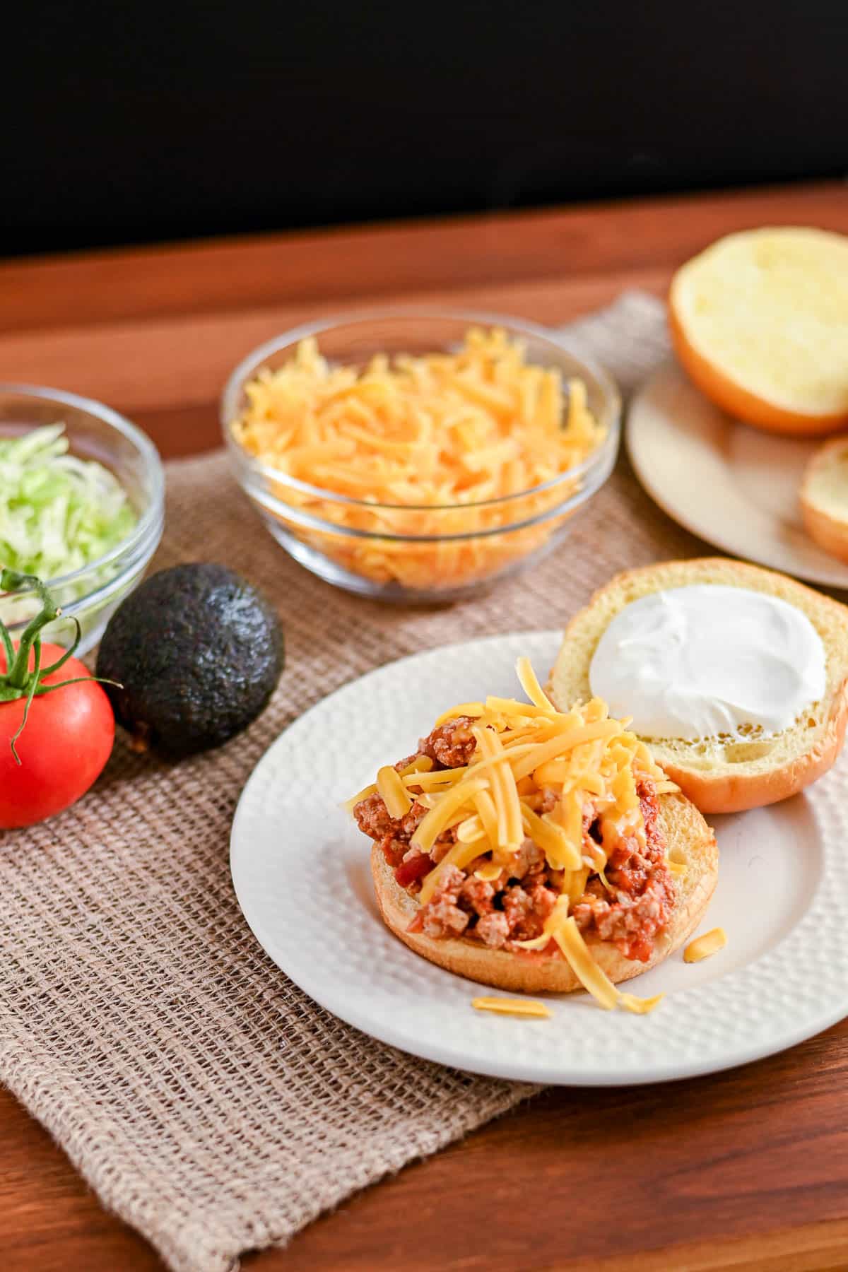 Open Taco Sloppy Joes sandwich on a white plate, topped with cheddar cheese, beside lettuce, avocado, and tomato—perfect for adding to your list of easy weeknight recipes.