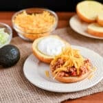 Open sandwich bun with ground meat, sour cream, and shredded cheese on a plate—perfect for Taco Sloppy Joes recipes—with fresh toppings in the background.