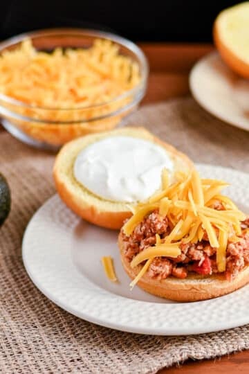 Open sandwich bun with ground meat, sour cream, and shredded cheese on a plate-perfect for Taco Sloppy Joes recipes-with fresh toppings in the background.