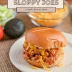 A Taco Sloppy Joes sandwich on a plate, topped with shredded cheese, avocado, and tomato in the background—perfect for quick weeknight recipes.