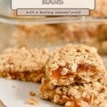 Three apricot bars with a crumbly oatmeal crust are stacked on parchment paper, reminiscent of classic Apricot Oatmeal Bars, with more bars in the background.