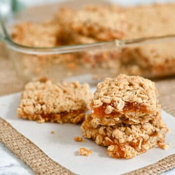 Two Apricot Bars with oat crumble and jam filling are stacked on parchment, with more bars in a glass dish in the background.
