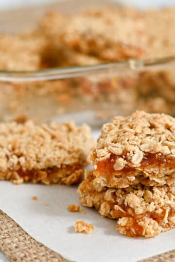 Two stacked Raspberry Oatmeal Bars sit on parchment, with more bars in a glass dish in the background.