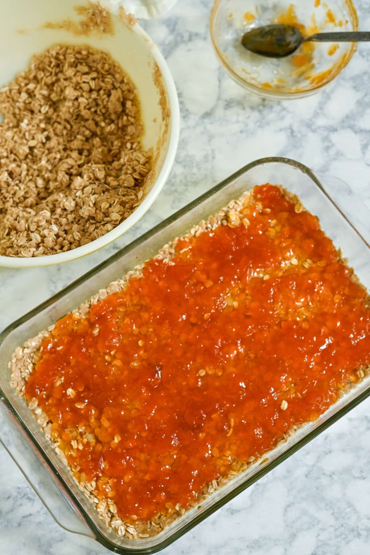 Glass dish with oat crumble base and orange jam, reminiscent of homemade Apricot Bars, next to a bowl of oat mixture and a spoon on a marble surface.