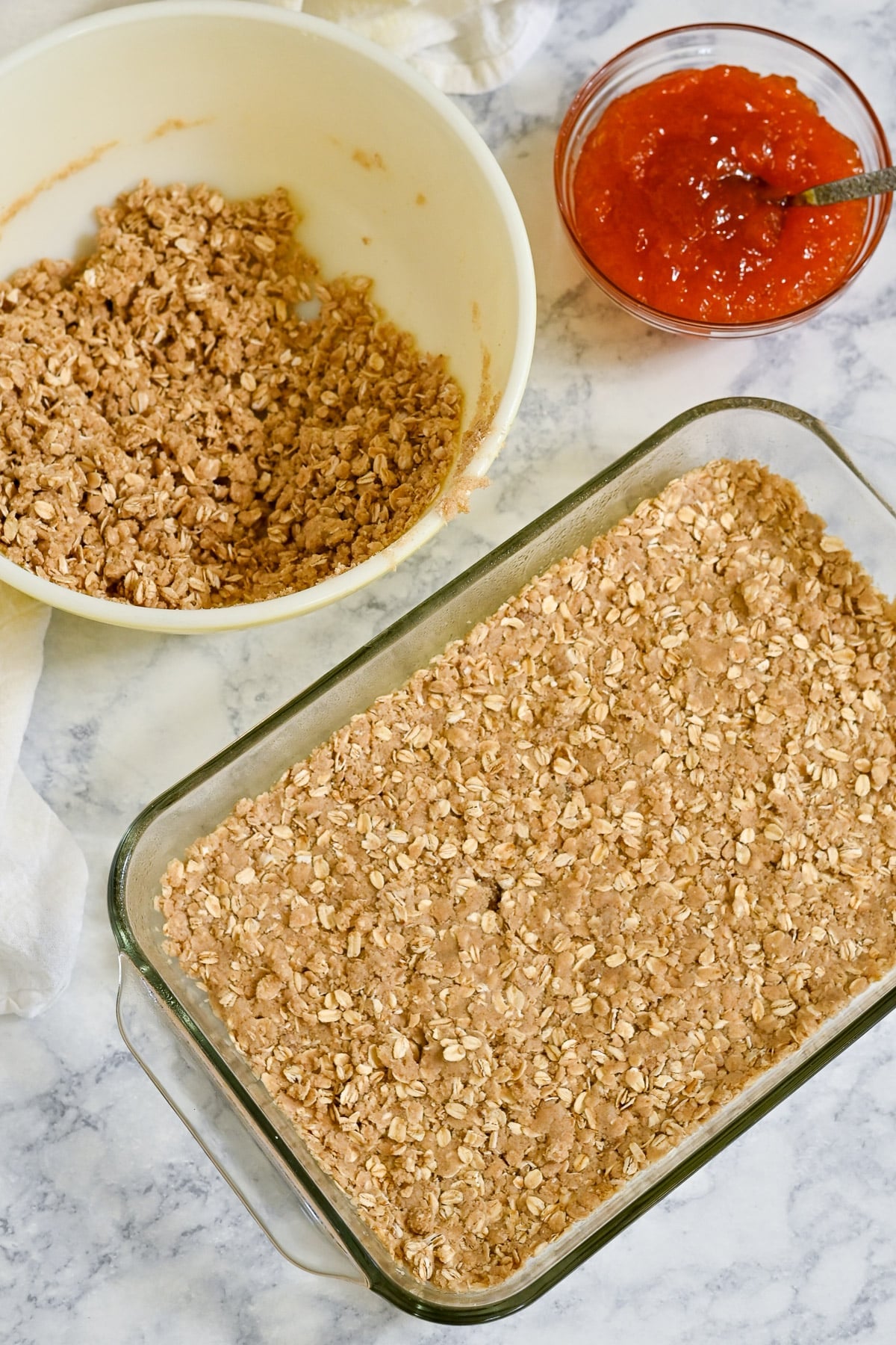 A baking dish with oat mixture for Apricot Bars, a mixing bowl with oats, and a bowl of red jam on a marble counter.