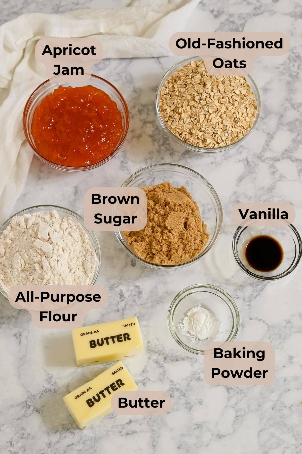 Seven labeled bowls and sticks with oats, flour, brown sugar, vanilla, jam, baking powder, and butter on marble-perfect ingredients for making irresistible Apricot Bars.