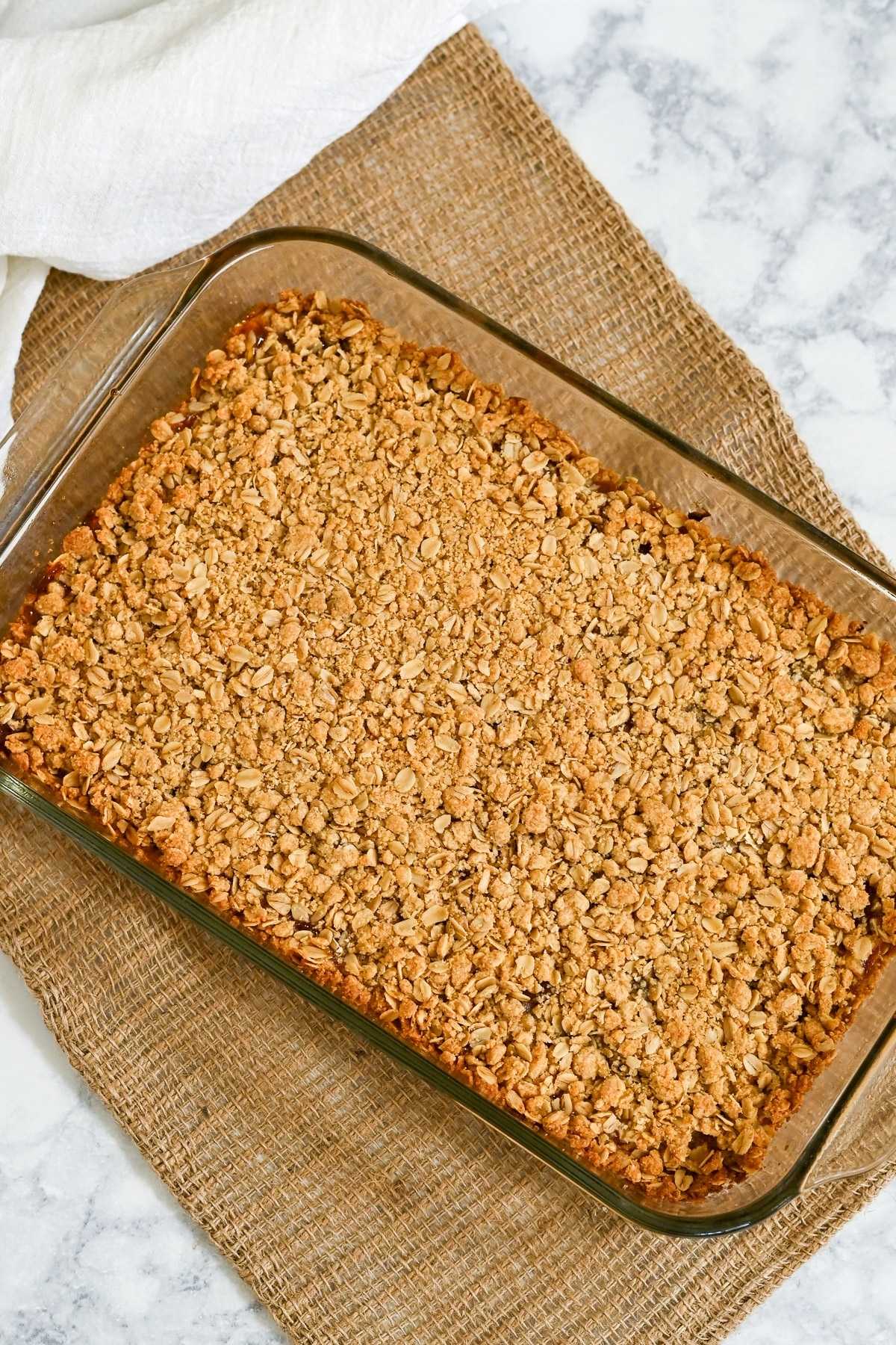 A glass baking dish filled with golden baked oat crumble, reminiscent of homemade Apricot Bars, sits on a burlap mat and marble surface.