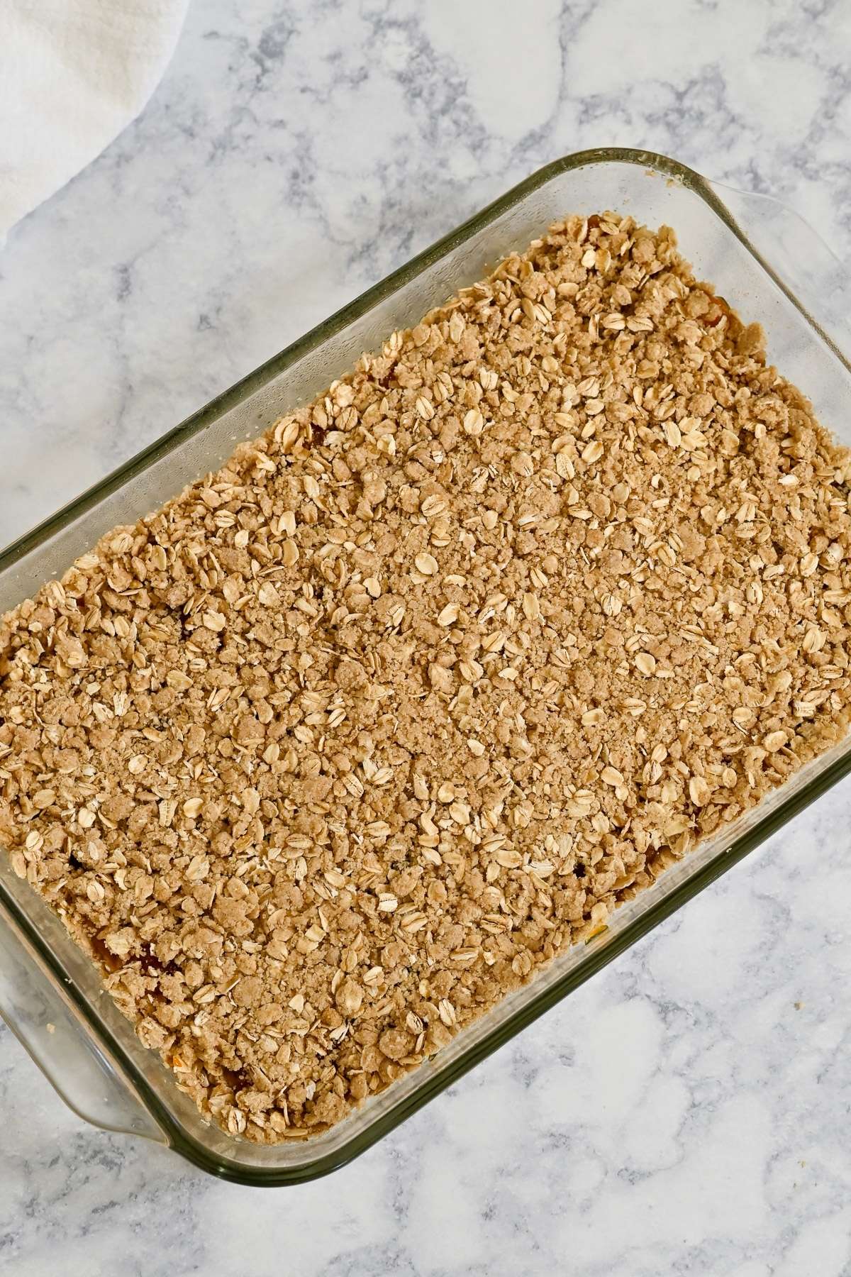 A glass baking dish filled with golden baked oat crumble, reminiscent of classic Apricot Bars, sits on a marble countertop.