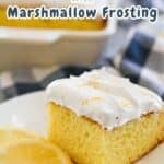 A slice of lemonade cake with marshmallow frosting on a white plate, garnished with lemon slices. The background shows a larger dish of the lemonade cake marshmallow frosting. Text reads "Lemonade Cake with Marshmallow Frosting.