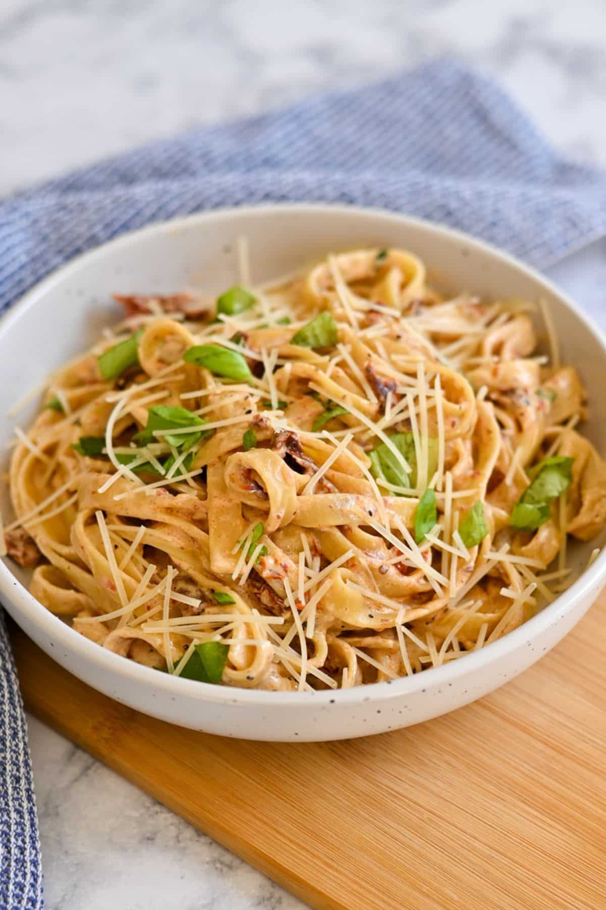 A bowl of Creamy Sun-Dried Tomato Pasta topped with grated cheese and fresh basil sits on a wooden board, its rich sauce enhanced by a touch of cream cheese.