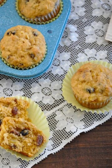 Muffins with raisins in and out of a blue muffin tray, one split in half on a lace tablecloth next to an old handwritten note. The scene evokes the charm of homemade Pineapple Nut Muffins, bringing warmth and nostalgia to mind.