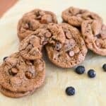 Blueberry Chocolate Chip Cookies rest on parchment paper, with some cookies stacked and one featuring a tempting bite. Scattered blueberries and rich chocolate chips make these treats irresistible.
